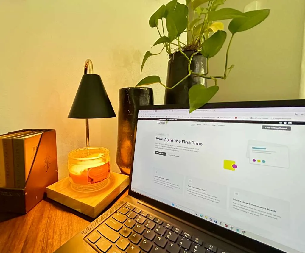 A laptop on a wooden desk next to a lit candle, books, and a thriving Pothos plant, showing a flexible work-from-home setup.